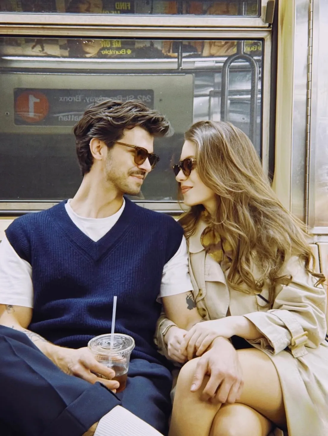 Romi and Niko sitting close together on a subway train, both in sunglasses, looking at each other.